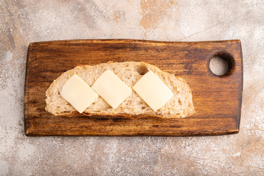 Slice of homemade bread with butter on cutting board on brown concrete, close up, top view.