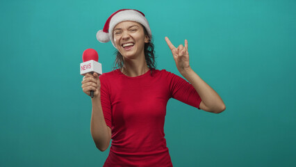 Fototapeta premium Woman wearing santa hat and red shirt holds a news microphone and makes rock hand sign in turquoise studio; festive cheer.