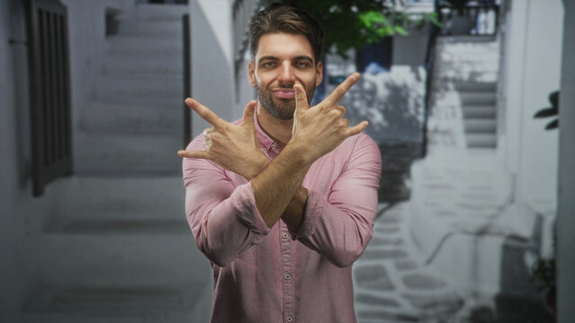 Man with beard in pink shirt flashing rock horns with hands and teeth bared while smiling on a narrow sunlit street; playful joy.