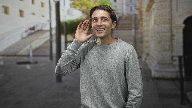 Man smiling and giving thumbs up with right hand on a street near a stone arch and planter; approval gesture.