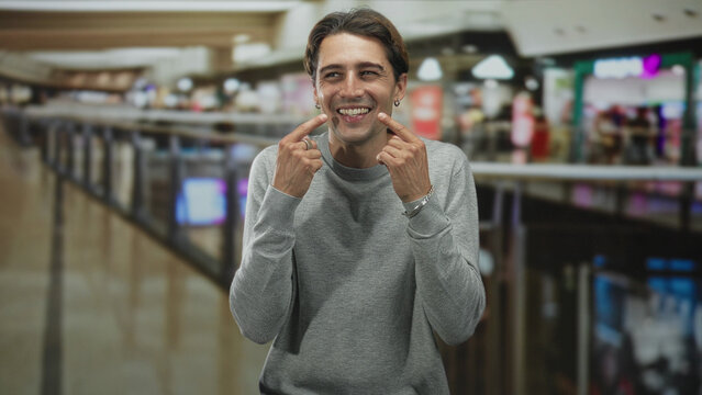 Man points index fingers to cheeks smiling in a busy mall building with metal railing, shops and blurred shoppers behind him, casual grey crewneck and jewelry; playful joy.