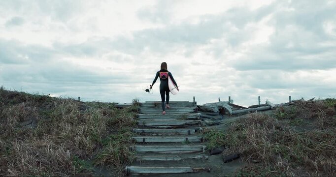 Back, woman and surfer at beach, walking up stairs and ready for fitness with wet suit, sports and surfing in nature. Steps, exercise and surfboard, ocean with professional or hobby with challenge
