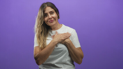 Obraz premium Woman with hands over chest wearing a white t shirt in a purple studio, smiling with eyes closed; gratitude warmth.