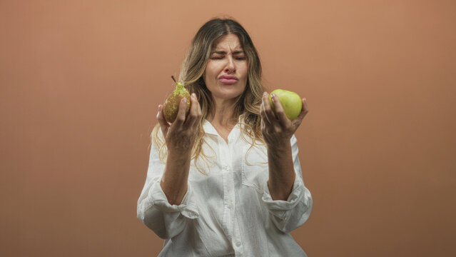 Woman holding a pear and green apple in hands while grimacing with pursed lips in a studio; taste decision disgust.