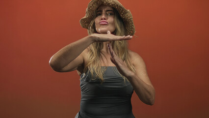 Fototapeta premium Woman makes time out gesture with palms horizontal and visible hands near chest, wearing straw hat and sleeveless top against orange backdrop in studio; confidence pause.