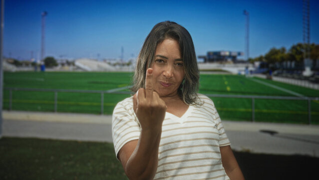 Hispanic woman showing middle finger to camera on a sunny athletic field with bleachers and fencing building; defiance.