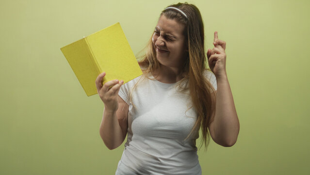 Woman holding yellow book close to face with fingers crossed, eyes closed and slight smile in studio; hopeful anticipation.