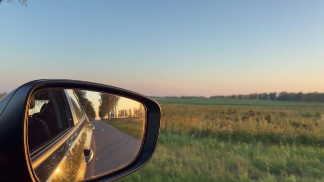 Car side mirror reflecting road and birch trees while driving past green fields at sunset. Peaceful travel and summer road trips through the countryside.