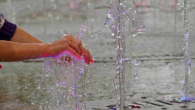 Little girl plays with hands with jets of a fountain in a city park.