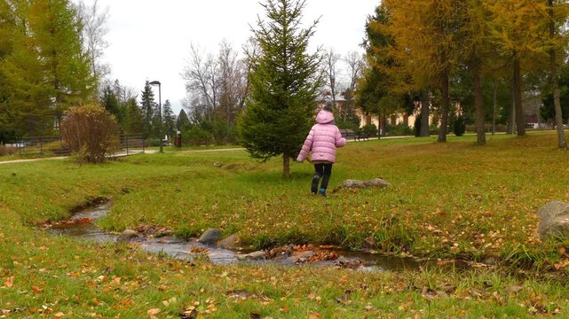 beautiful girl in a red jacket jumps over a small stream in the autumn city park