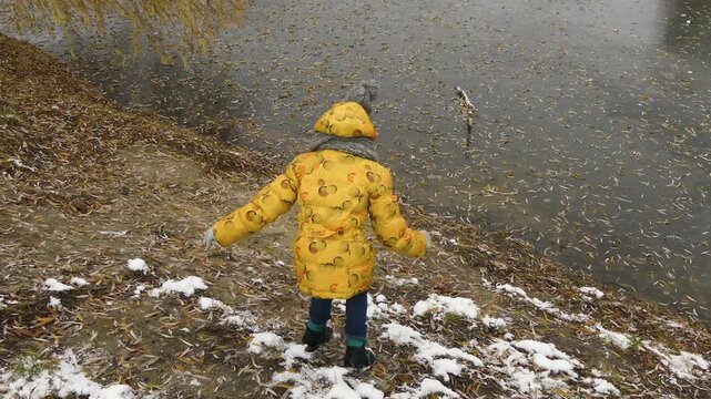 Small beautiful girl in yellow jacket goes to frozen pond in old city park.