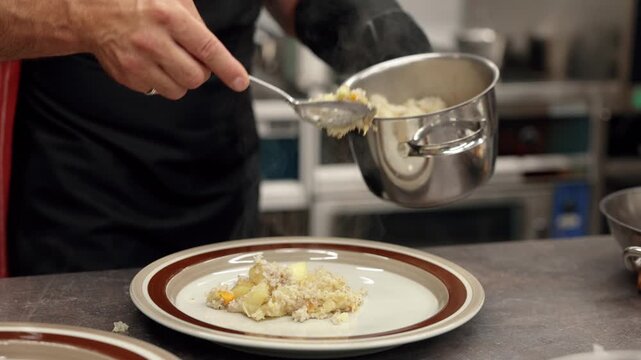 Close-up of a chef spooning a hot cooked dish from a stainless steel pot onto a ceramic plate in a professional kitchen, with visible steam