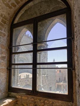 Horizontal composition featuring stone embrasures, wooden window panes, and a framed perspective of Erice's rooftops from inside a medieval tower.