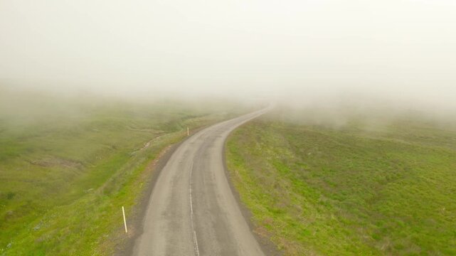 An aerial view of a winding asphalt road through a green, grassy landscape. The road curves and disappears into a thick, white fog that obscures the horizon.
