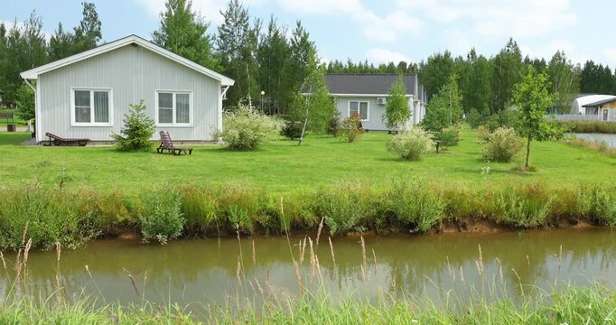 Modern glamping site with cozy waterfront cabins next to scenic pond. Luxury eco tourism resort featuring minimalist wooden houses on green lake shore. Peaceful countryside vacation destination surrou