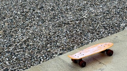 Orange skateboard resting on a concrete path beside gravel, stationary outdoor urban scene, youth leisure, board sport. © evgeniya