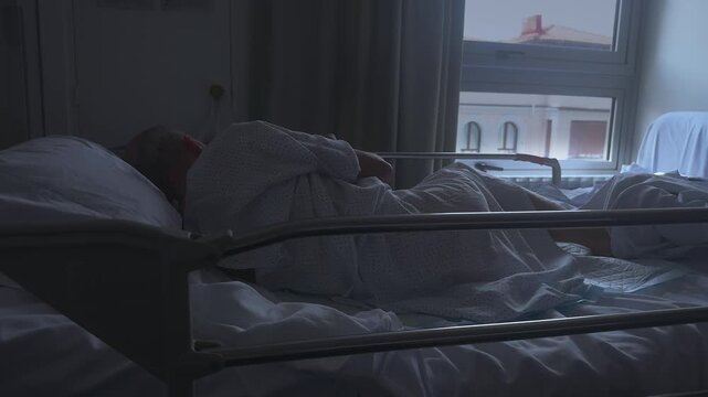 An elderly patient lying in a hospital bed, alone in a dimly lit room.