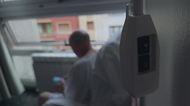 A lonely hospital patient sits alone fidgeting while looking out of the hospital window.