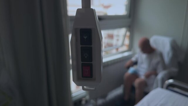 A hospital patient sits alone in the corner of a hospital room with the call button hanging in the foreground. Blurred anonymous male looking out the window.