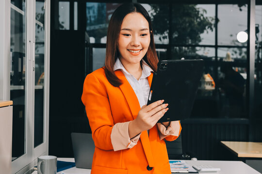 Smiling asian businesswoman talking on phone and using laptop while working at office desk