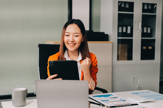 Smiling asian businesswoman talking on phone and using laptop while working at office desk