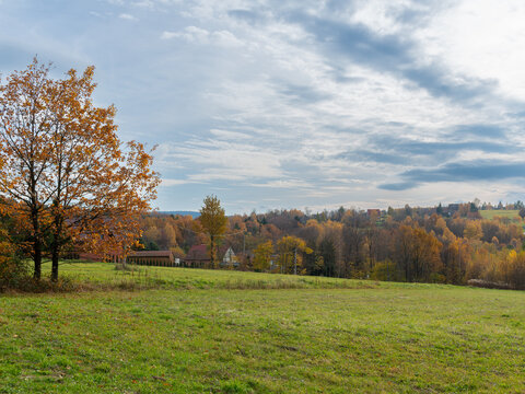 Open green meadow overlooks scattered rural houses, wooded hills and colorful autumn trees beneath a layered cloudy sky. Spacious countryside landscape suitable for rural tourism, seasonal nature, vil