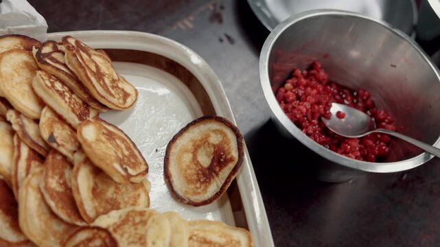 Golden mini pancakes rest on a plate beside a stainless steel bowl filled with chunky red berry compote and a spoon on a dark kitchen work surface
