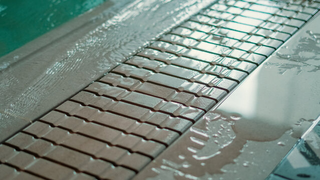 A close-up view of water overflowing the edge of an indoor swimming pool onto a wet tiled drainage grate and patterned floor
