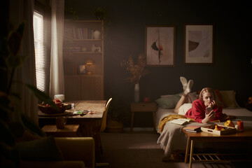 Young Caucasian woman lying on bed and using smartphone in cozy bedroom. Image represents digital lifestyle and remote leisure, suitable for mobile application marketing © DragonImages