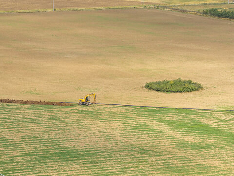 Yellow excavator digging a trench for pipeline installation in vast rural agricultural field under bright daylight, highlighting infrastructure development and earthmoving work.
