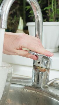 Close up of hand turning chrome faucet handle to control water. Water flows into glass and sink creating ripples. Background includes white countertop green plants and ceramic vase