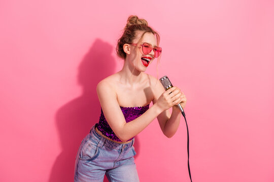 Happy young woman in a stylish outfit singing joyfully into a microphone against a vibrant pink background