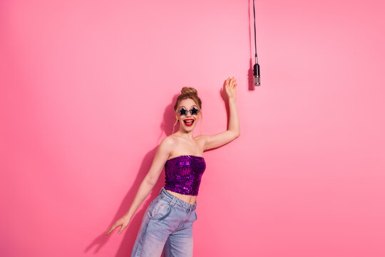 Trendy young woman in stylish attire enjoying music with retro microphone against vibrant pink background