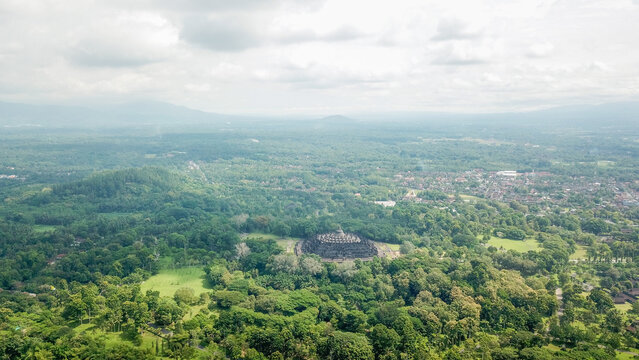 Indonesia - 23 February 2023: Aerial view of the ancient Borobudur Temple surrounded by lush green tropical forests and distant mountains under a cloudy sky.