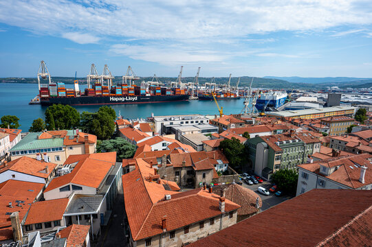 Koper, Slovenia - August 07, 2026: Panoramic high-angle view of red rooftops in the old town with cargo ships and port cranes in the background.