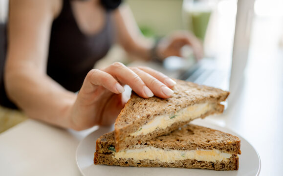 Person holds egg salad sandwich on whole wheat bread while working at desk during daytime