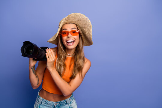 Young chic woman photographer with straw hat wearing orange top smiles holding a camera against a purple background