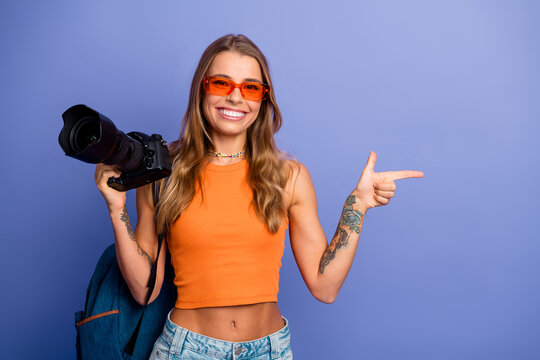 Young chic photographer with orange top camera in hand pointing to the side smiling for lifestyle fashion image