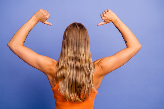 Back view of a young woman with blonde hair wearing an orange singlet showing thumbs down against a blue background