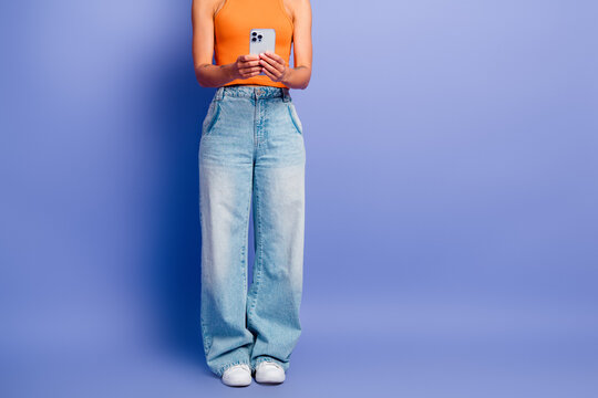 Young stylish woman in orange top and wide jeans using smartphone against blue background
