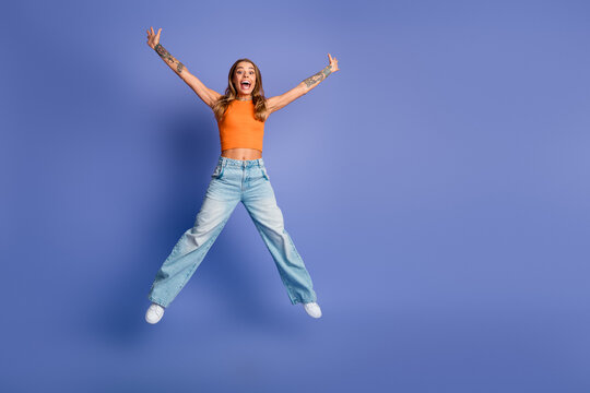 Young chic woman jumps in orange top and jeans against purple background expressing joy and energy