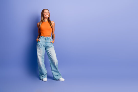 Young stylish woman wearing orange singlet and wide jeans stands against a blue background smiling and posing for fashion lifestyle shoot