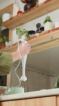 Person in green sleeves uses white immersion blender. Background features wooden shelves with potted plants. Glass jar filled with water is visible near mixing containers