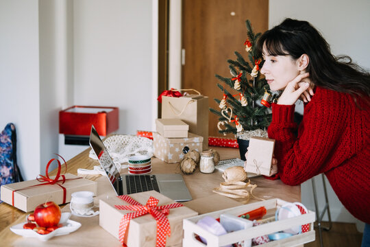 Woman leans on table holding kraft parcel while looking at laptop among wrapped gifts. Small business christmas orders, packing handmade gifts christmas, online shop christmas preparation