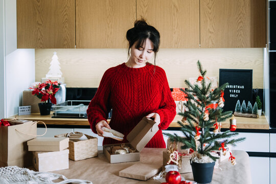 Woman holds wooden brush over open kraft box in kitchen near CHRISTMAS VIBES ONLY sign. Small business assembling christmas gift set, handmade spa gift box preparation, artisan business product assemb
