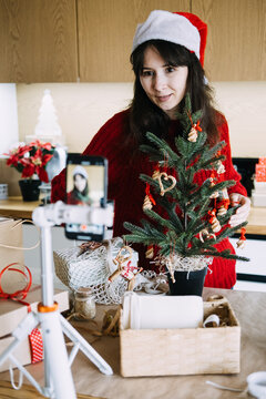 Woman in Santa hat decorates small potted tree while phone on tripod records her in kitchen. Filming christmas vlog phone, content creator holiday video, recording festive video content