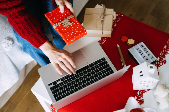 Hands hold wrapped gift above laptop keyboard on table with parcels and pen. AI christmas gift recommendation, chatbot shopping assistant, personalized gift suggestions online