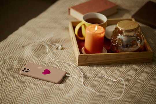 Smartphone with earphones lying on bed next to wooden tray with teapot and burning candle, representing cozy relaxation. Ideal for mental wellbeing and digital detox campaigns