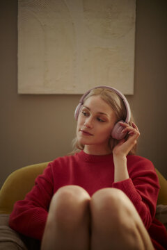Vertical image of young Caucasian woman wearing headphones, listening to music or podcast while sitting in armchair. Concept of mental wellbeing, perfect for audio app marketing