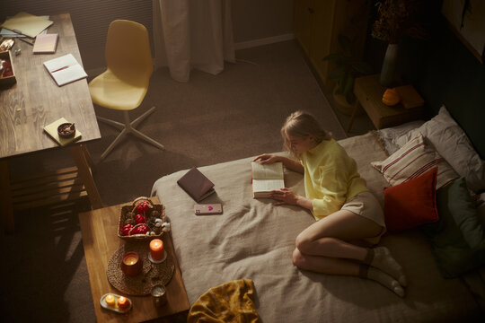 Young Caucasian woman lying on bed and reading book in cozy bedroom. High angle view showing self care and mental wellbeing concept, perfect for lifestyle marketing